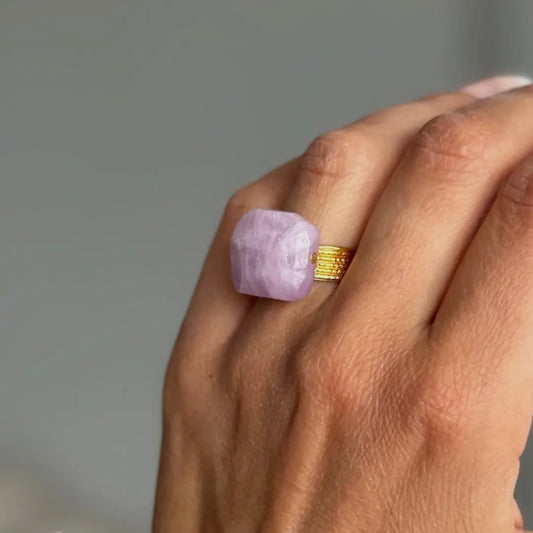 Hand wearing a ring with a purple gemstone and gold band on a neutral background